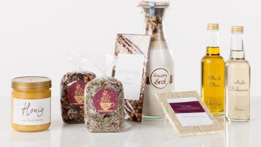 Ένα σετ προϊόντων delicatessen με ταιριαστές ετικέτες: ένα βάζο μέλι, σακουλάκια με βότανα και γκρανόλα, μπουκάλια με ελαιόλαδο και βαλσάμικο ξύδι, και μια πλάκα σοκολάτας.