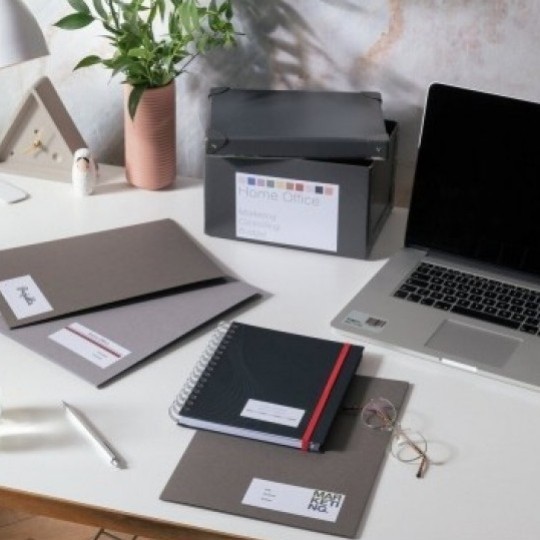 Ένα οργανωμένο γραφείο στο σπίτι με laptop, φακέλους, ένα σημειωματάριο και ένα κουτί αρχειοθέτησης, πάνω στα οποία έχουν κολληθεί λευκές ετικέτες οργάνωσης με κείμενα όπως "Home Office" και "MARKETING".  Zapytaj  Wyjaśnij