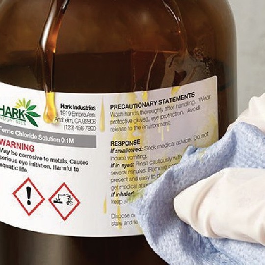 Χημικά ανθεκτική ετικέτα με την αναγραφή «Ferric Chloride Solution» και εικονογράμματα GHS σε ένα καφέ μπουκάλι, την οποία σκουπίζει ένα χέρι με γάντι χρησιμοποιώντας ένα πανί.