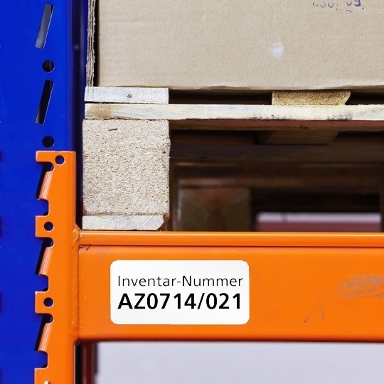 Λευκή ετικέτα απογραφής με την ένδειξη "Inventar-Nummer AZ0714/021", κολλημένη σε μια πορτοκαλί δοκό ραφιού αποθήκης, πάνω στην οποία βρίσκεται μια παλέτα με ένα χαρτοκιβώτιο.