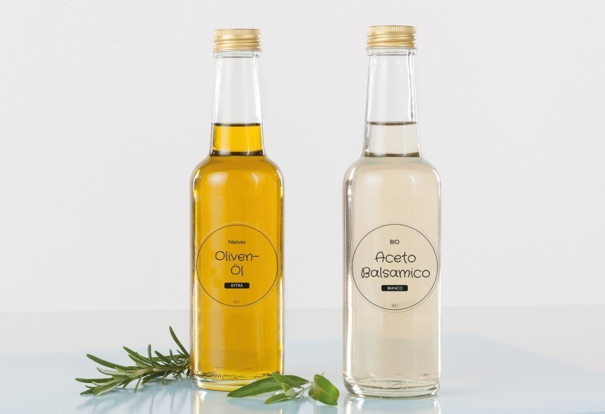 Δύο γυάλινα μπουκάλια: το ένα με έξτρα παρθένο ελαιόλαδο, το άλλο με λευκό βαλσάμικο ξύδι. Δίπλα βρίσκεται ένα κλαδάκι δεντρολίβανου. Οι ετικέτες είναι στρογγυλές και μινιμαλιστικές.