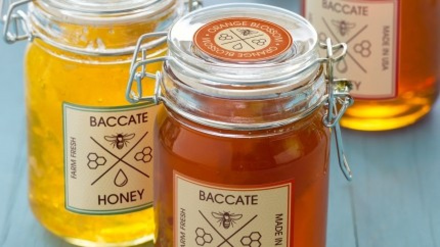 Δύο γυάλινα βάζα με μέλι 'Baccate' που φέρουν ετικέτες. Το βάζο στο προσκήνιο έχει ένα επιπλέον, στρογγυλό αυτοκόλλητο 'Orange Blossom' στο καπάκι.