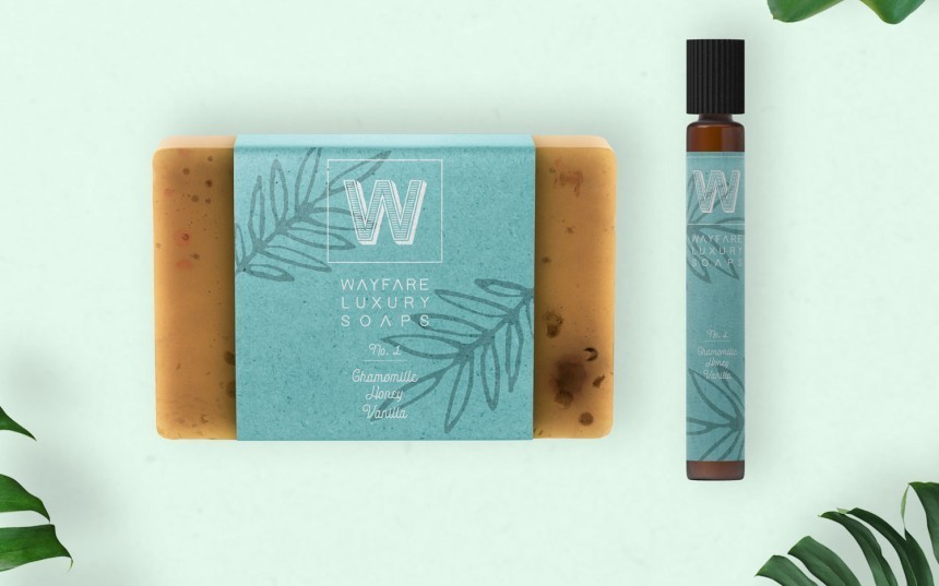 Κάτοψη μιας μπάρας χειροποίητου σαπουνιού και ενός μπουκαλιού roll-on. Τα προϊόντα έχουν ταιριαστές, τιρκουάζ ετικέτες με το λογότυπο 'W', την επιγραφή 'Wayfare Luxury Soaps' και ένα μοτίβο φύλλων.