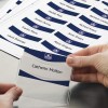 Η εικόνα δείχνει ένα χέρι να κρατάει ένα καρτελάκι με το όνομα 'Catherin Molton', που περιβάλλεται από ένα εκτυπωμένο φύλλο με πολλαπλά καρτελάκια ονομάτων στο παρασκήνιο.