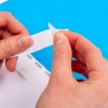 Η εικόνα δείχνει ένα κοντινό πλάνο από χέρια που ξεκολλάνε μία λωρίδα αυτοκόλλητων ετικετών από ένα υπόστρωμα. Το φόντο είναι ένα συμπαγές μπλε χρώμα.